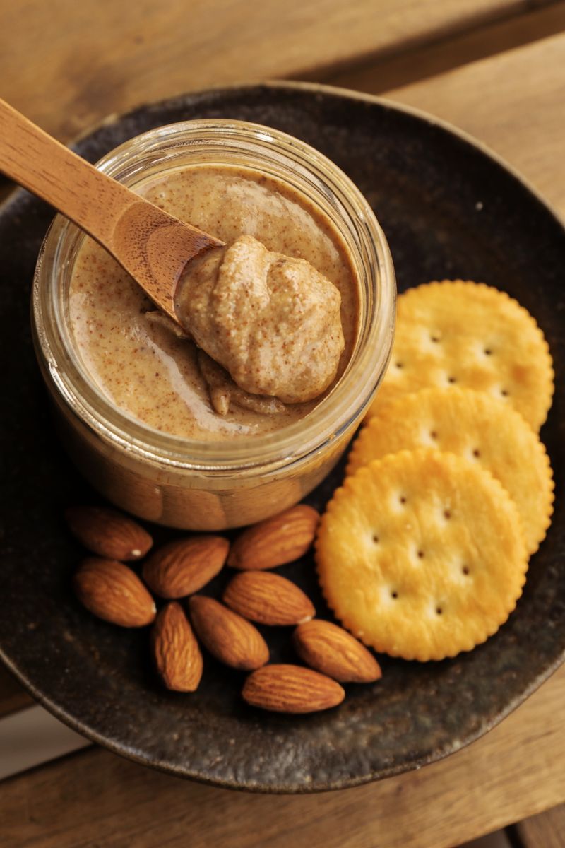 A jar of creamy almond butter with a wooden spoon, surrounded by almonds and crackers.